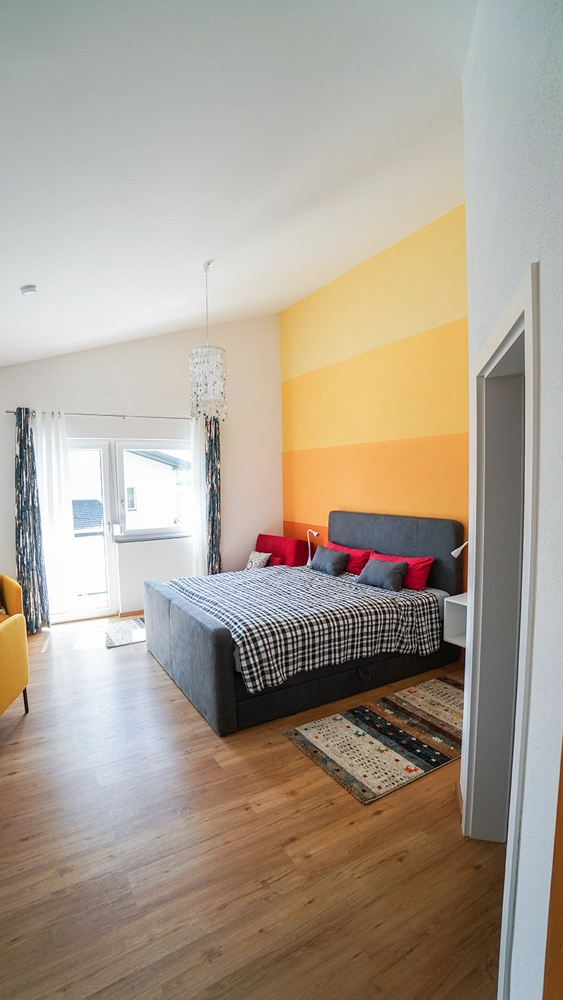 „Hahns Herberge“ – Innenansicht Schlafzimmer; Die Farben sind in verschiedenen Orangetönen und rot gehalten, aufgeräumtes, helles Ambiente.